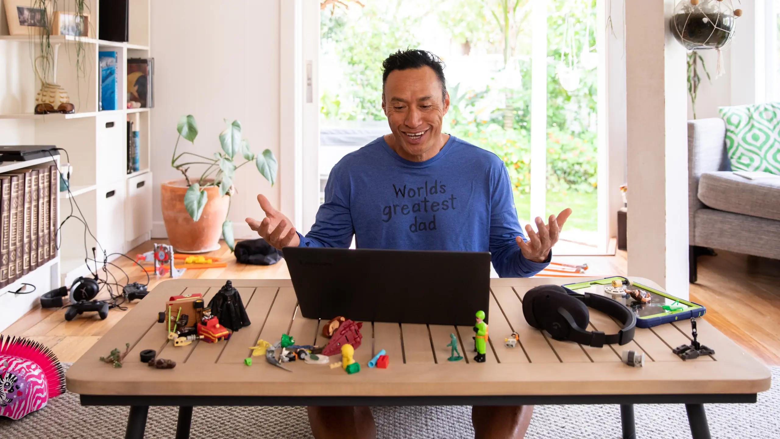Man at kitchen table looking amused at a laptop with kids toys on the table.
