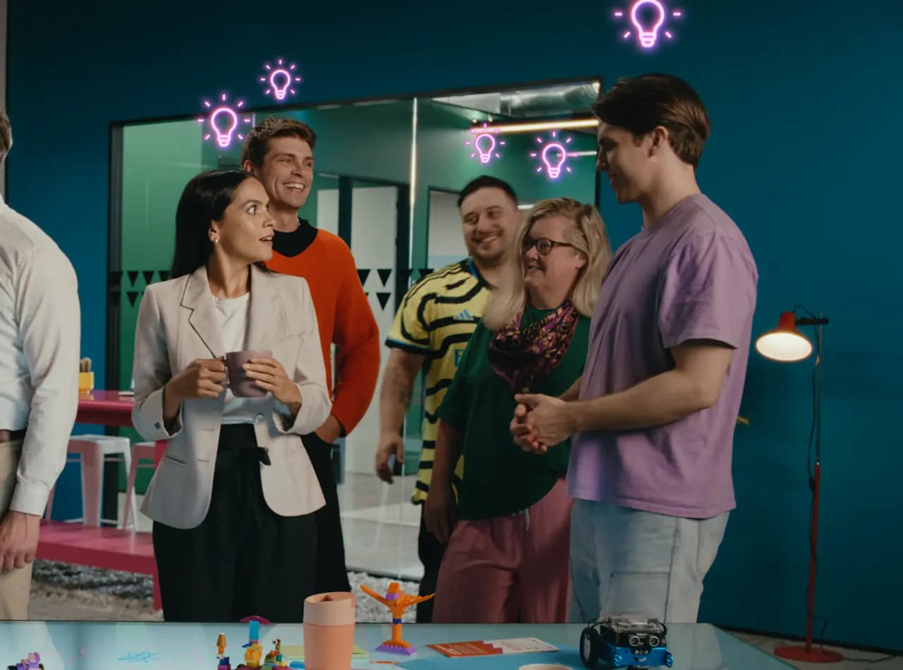 A group of five individuals in a bright office with a pink table and colorful decorations. Neon lightbulb icons appear above their heads, with scattered objects like toys and a mug on the table.