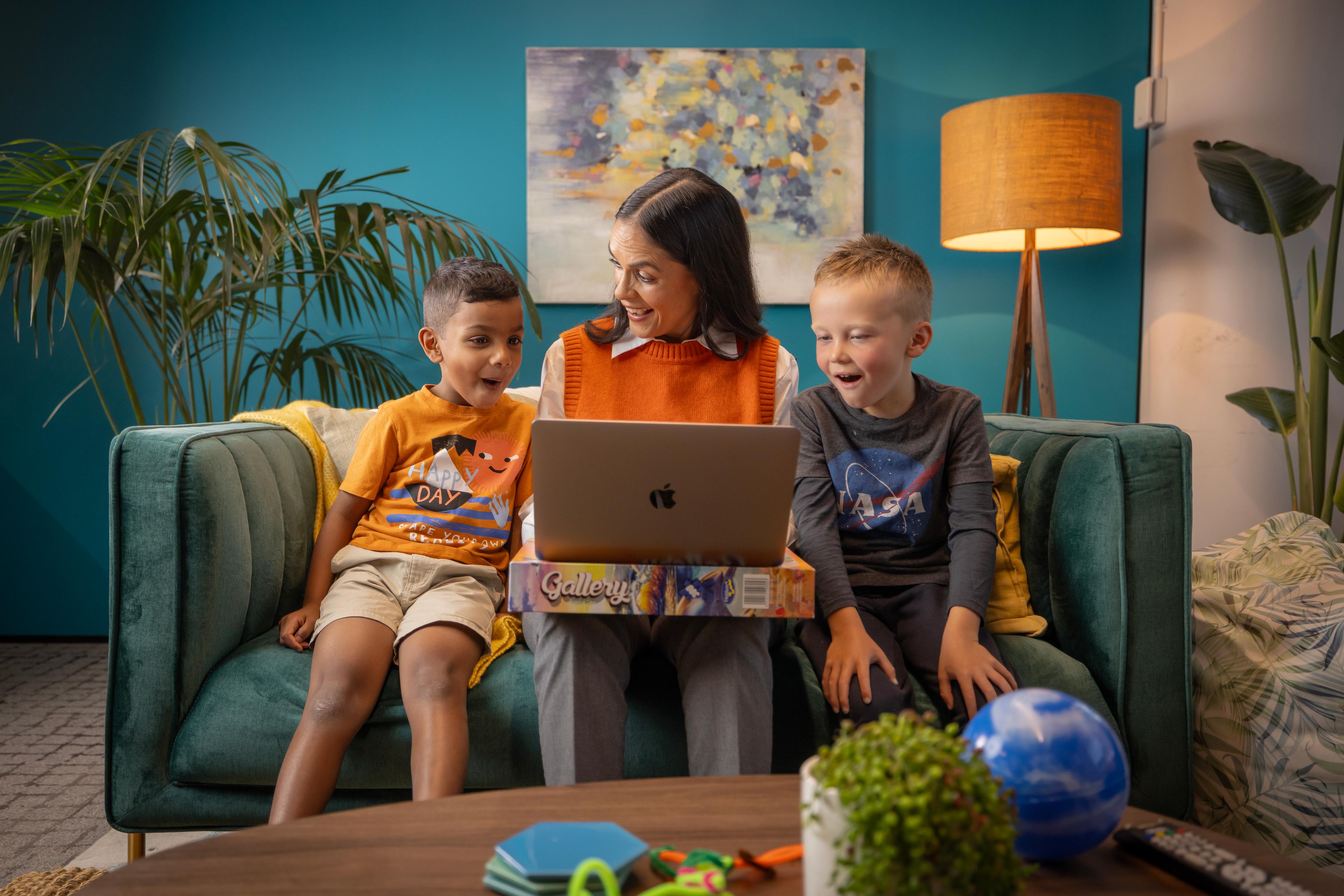 A woman and two boys smile while looking at a laptop on a teal sofa, surrounded by educational toys.