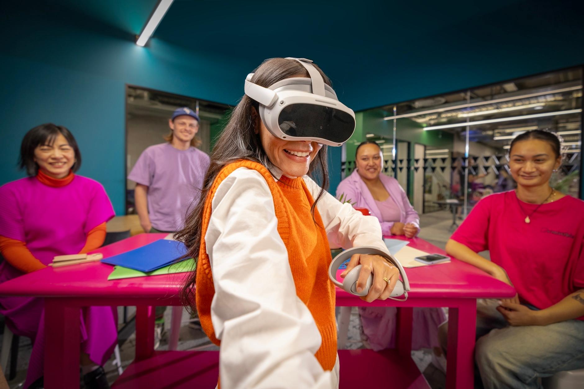 A woman in a VR headset smiles, surrounded by students and an instructor in a modern classroom.