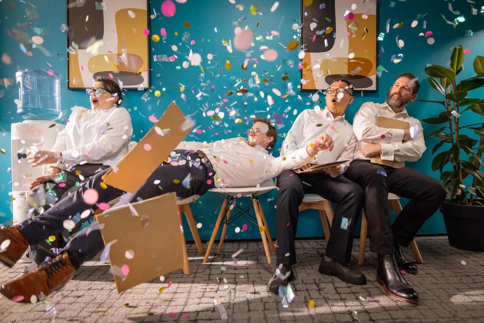 Four people in business attire get surprised as confetti bursts around them in a vibrant office lounge.