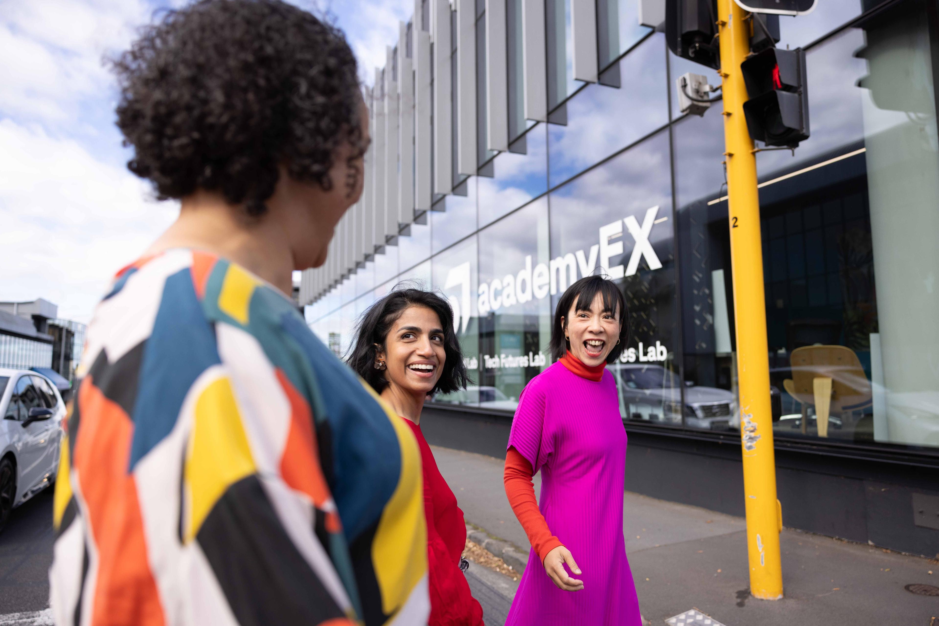 Three people are walking outside a modern building with a sign that reads "AcademyEX."