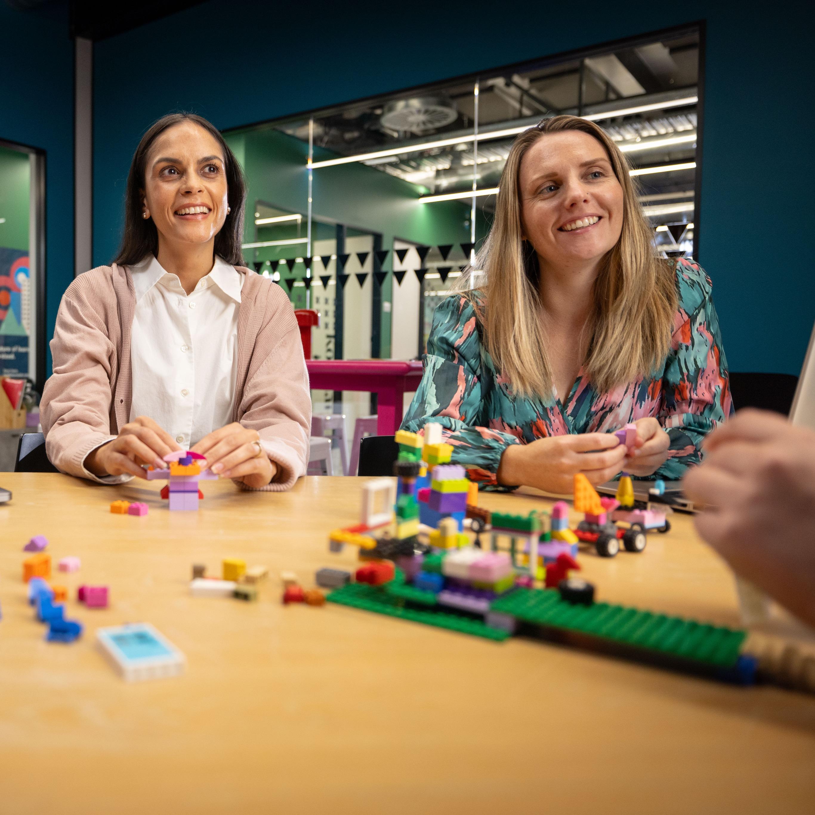 Two women smiling and building with LEGO bricks in a modern office with glass walls and vibrant decor. Collaborative and cheerful.