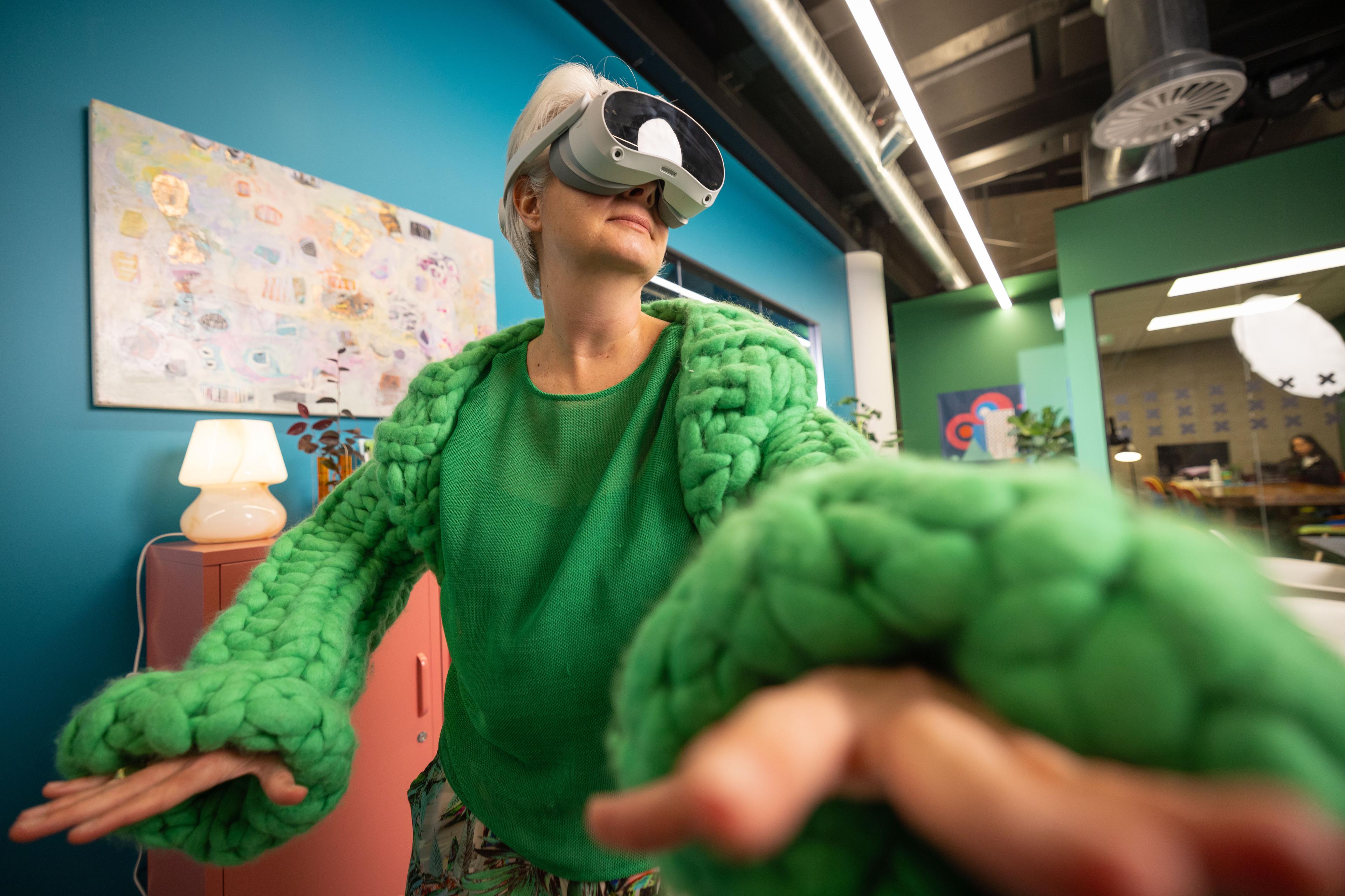 A person wearing a virtual reality headset and a bright green knit sweater extends their arm as if interacting with a virtual environment.