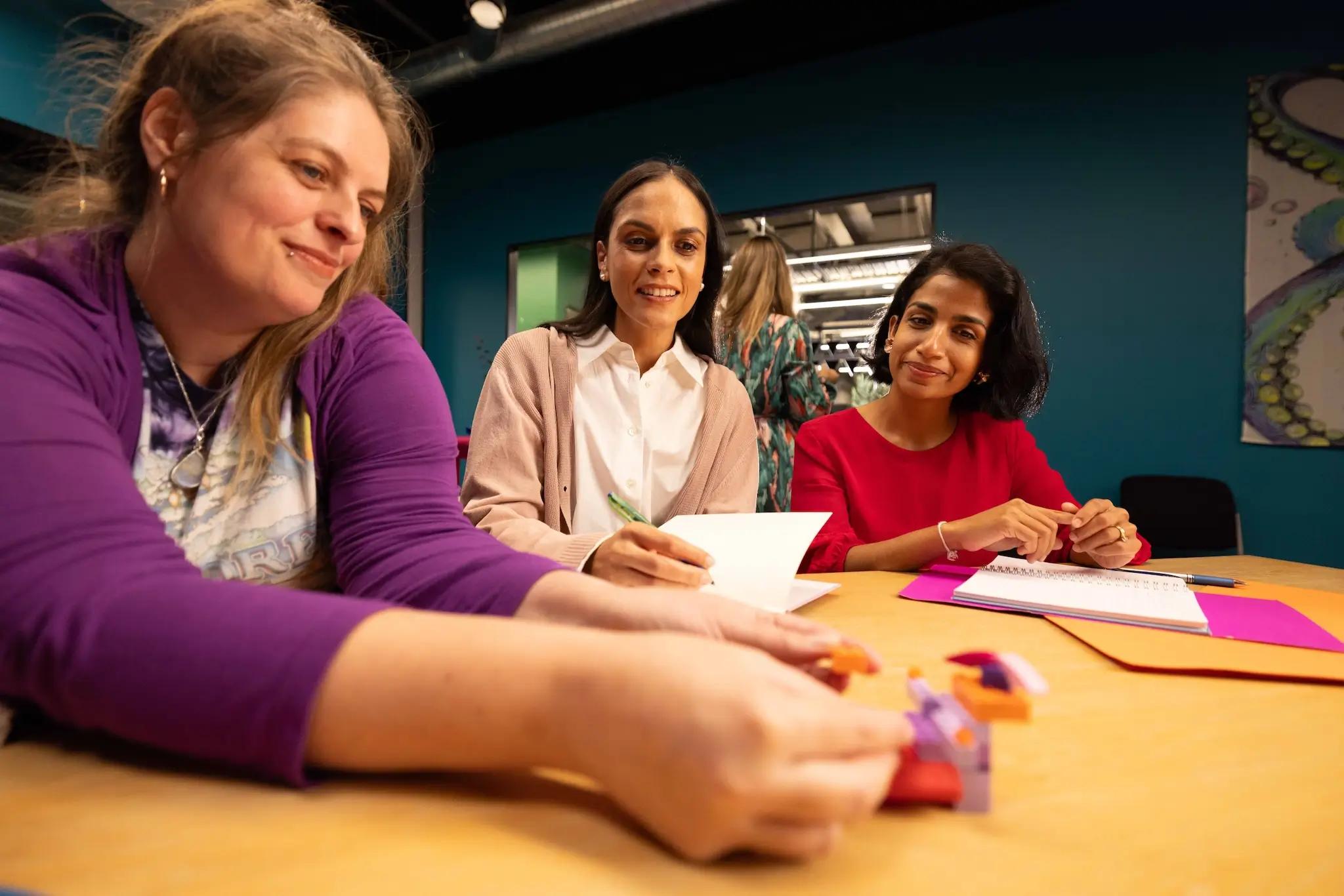 Three women collaborating at a colorful workshop table, with one assembling puzzle pieces.