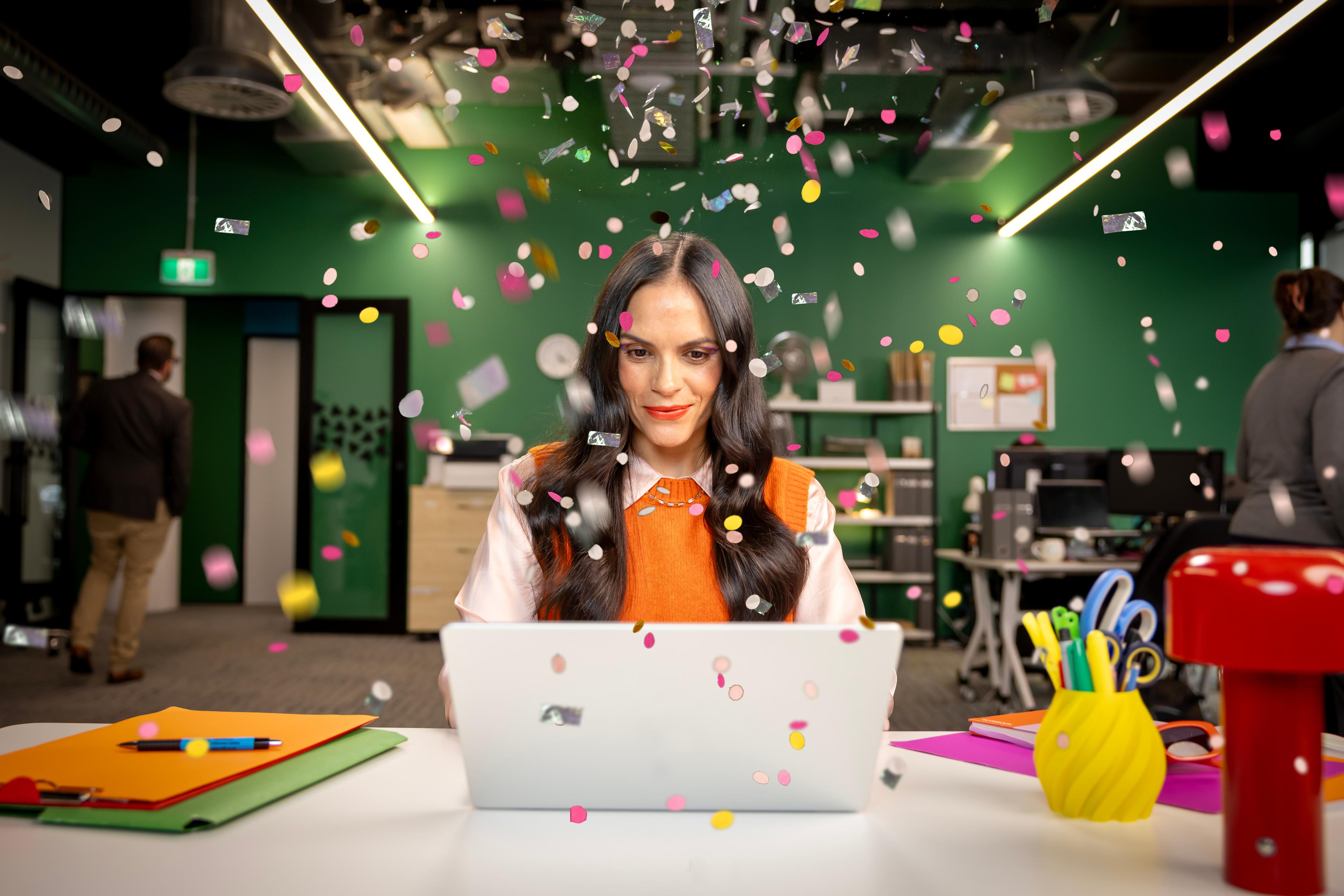 A woman at her desk, celebrating with confetti in an office with a green backdrop.