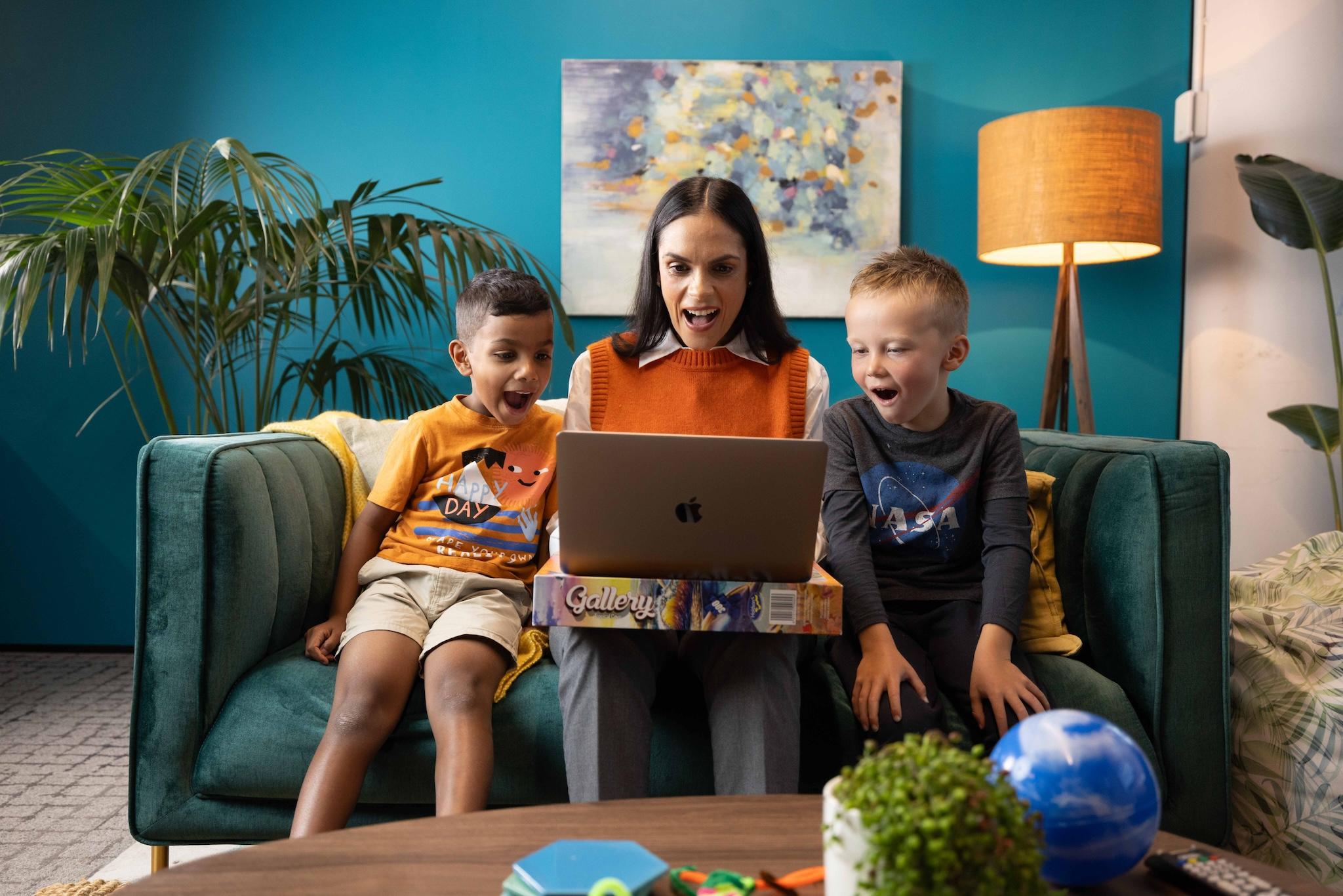 A woman and two boys smile in excitement while looking at a laptop on a teal sofa, surrounded by educational toys.