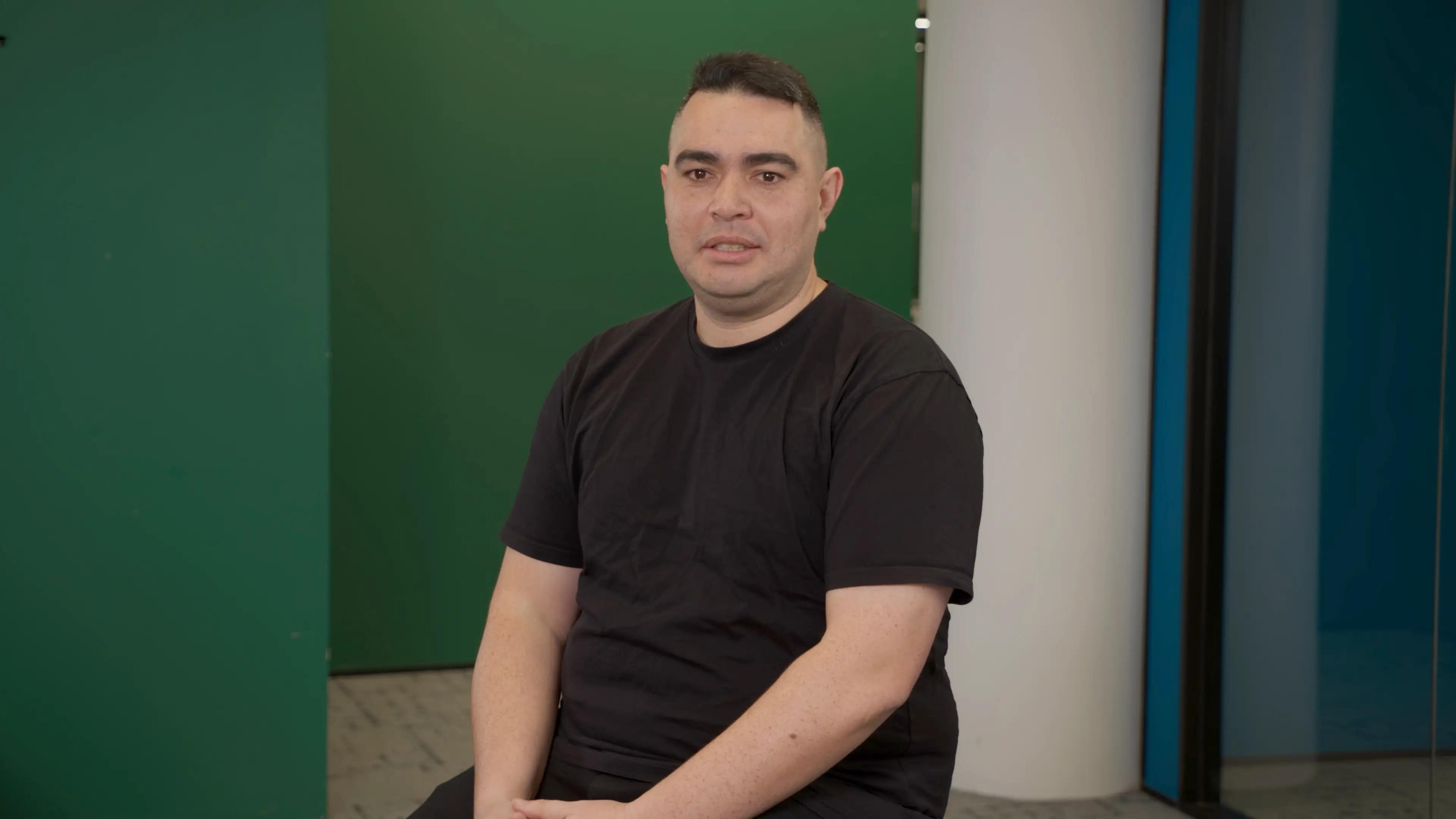 Joe Osborn seated against a plain green backdrop, dressed in a black t-shirt for his academyEX testimonial video.
