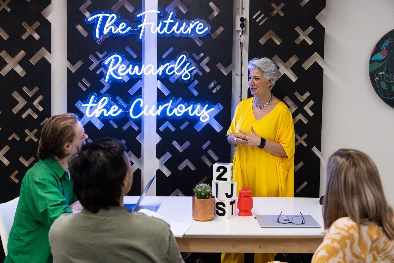 A woman in a yellow dress engages with her audience in a modern, neon-lit presentation about curiosity and the future