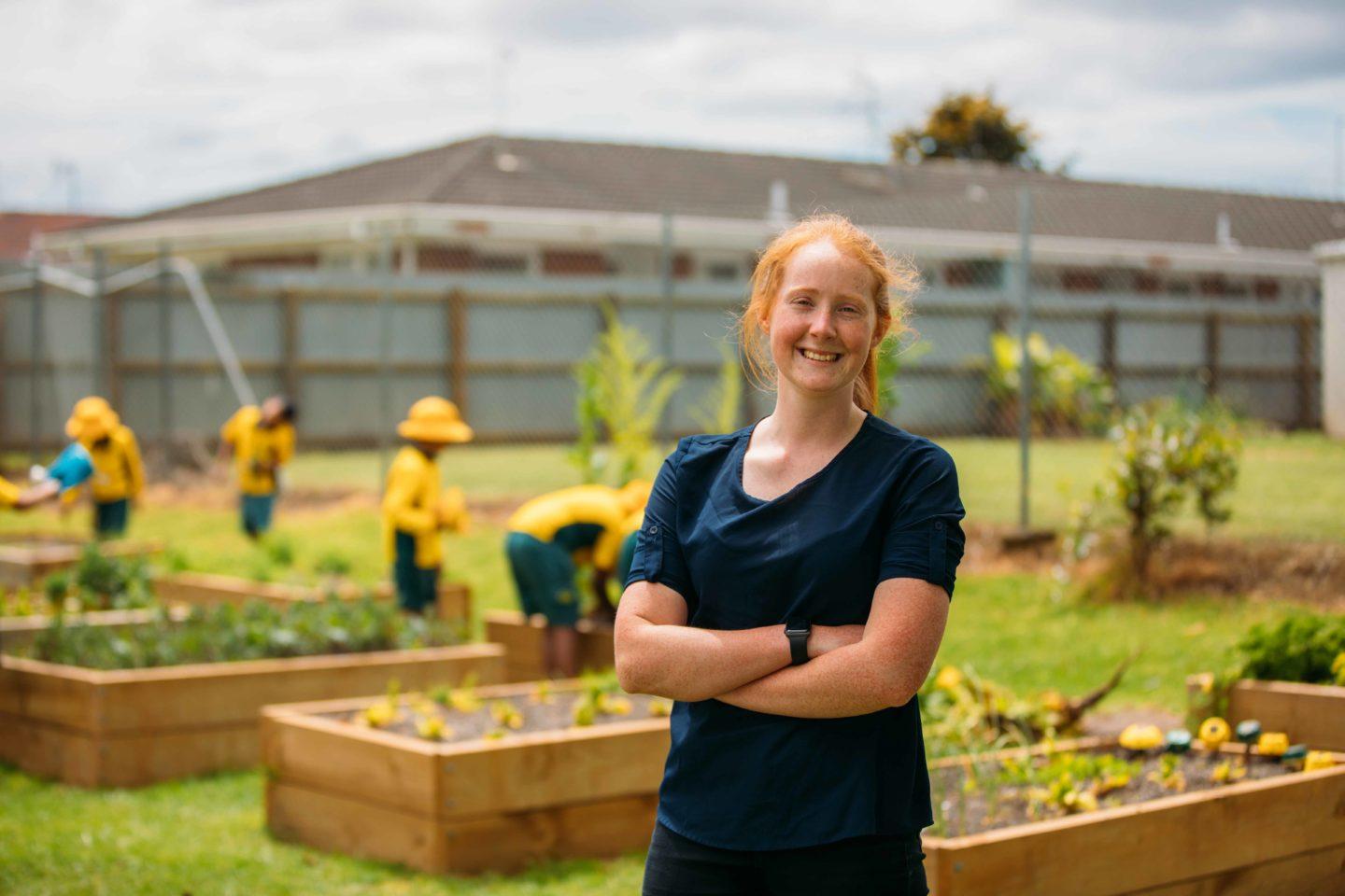 A teacher smiling with school students gardening in the background.